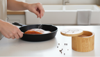 Person seasoning food in a pan on a kitchen counter with a bamboo salt box nearby.